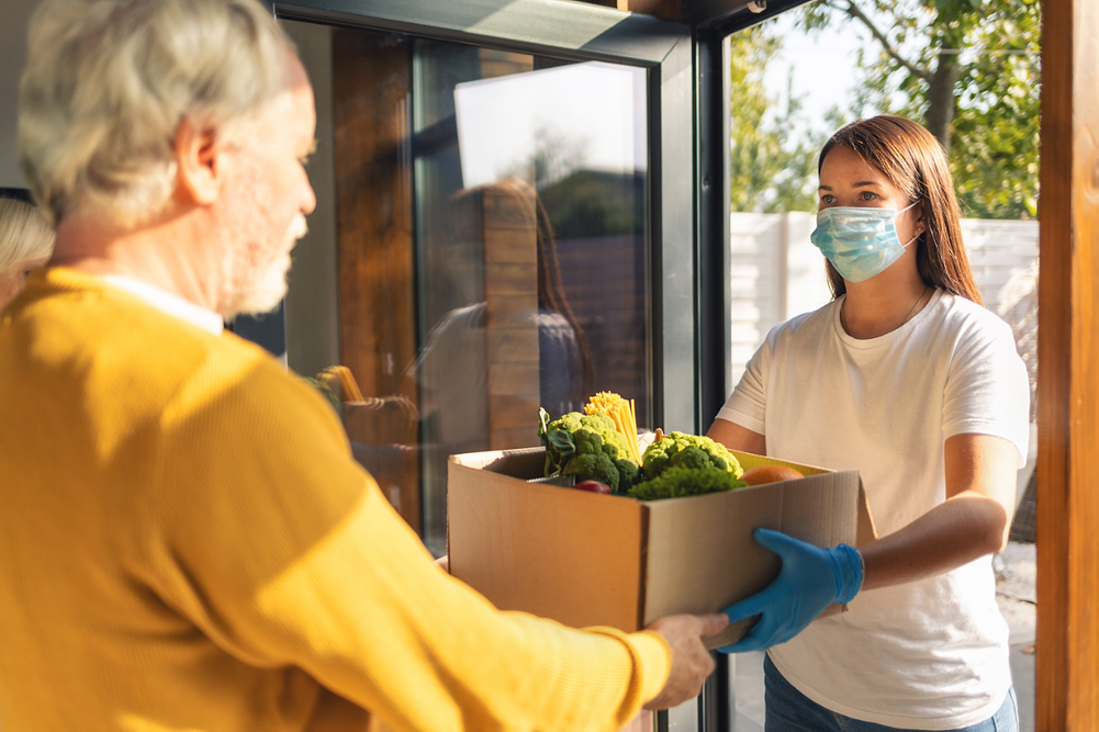 Young woman courier delivering purchases to elderly couple at doorway wearing mask and gloves