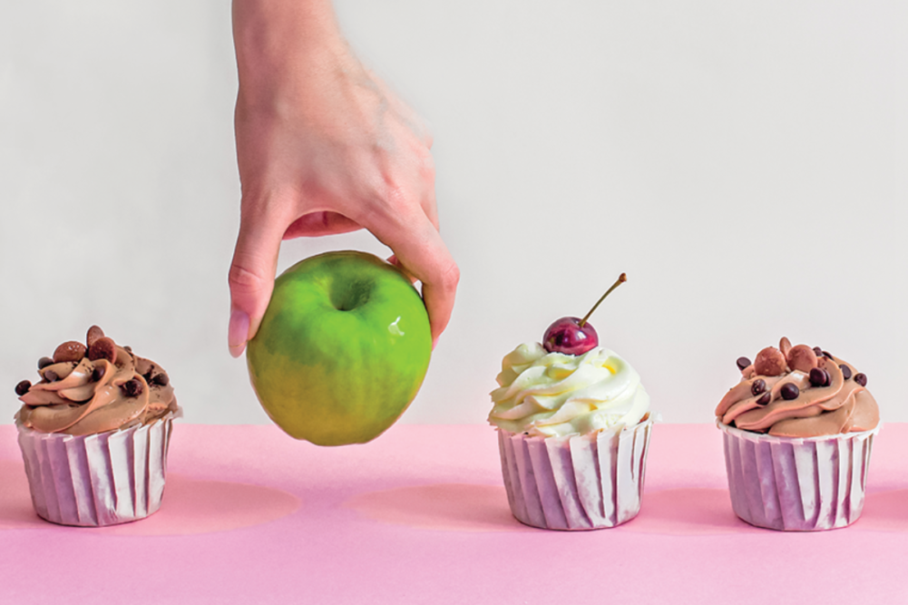 Hand grabbing an apple from a lineup of cupcakes
