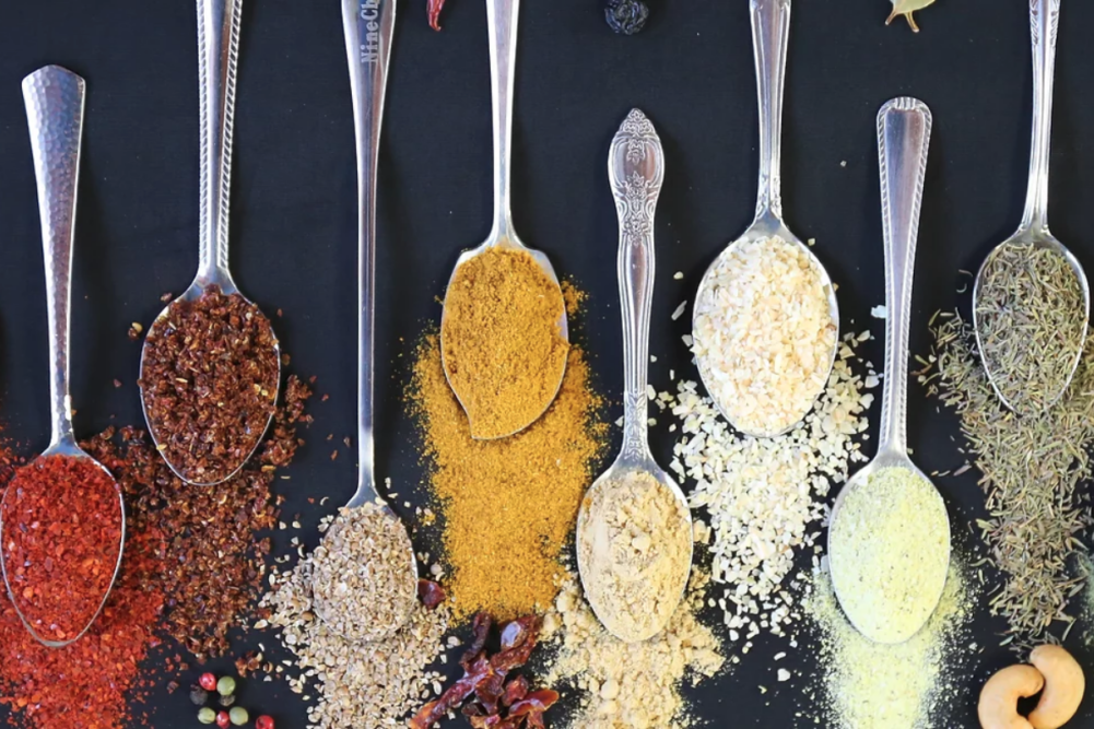 Row of spoons filled with spices and seasonings on blue background